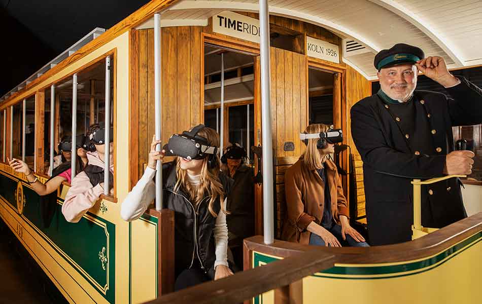 Fahrgäste mit VR-Brillen in historischer Straßenbahn, Schaffner tippt an Mütze.