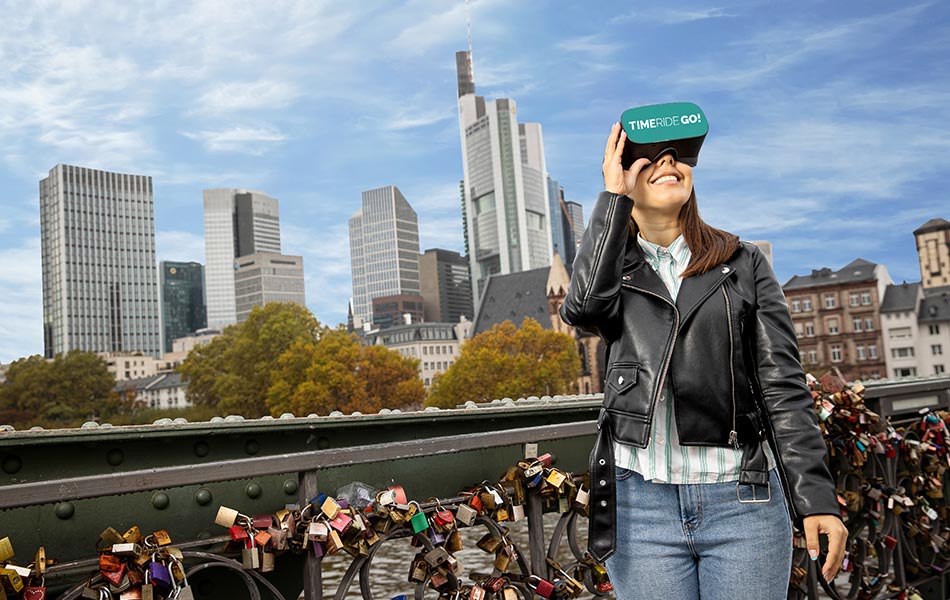 Frau mit VR-Brille vor moderner Frankfurter Skyline auf Brücke. Vorhängeschlösser hängen am Brückengeländer.