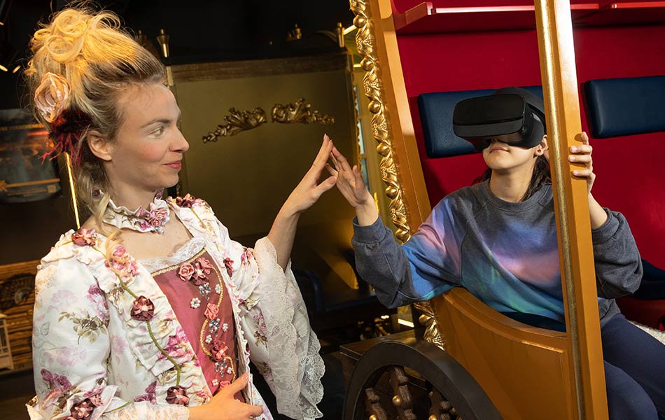 Dame in Barock-Kleid berührt die Hand eines Kindes das mit VR-Headset in einer goldenen Kutsche sitzt.