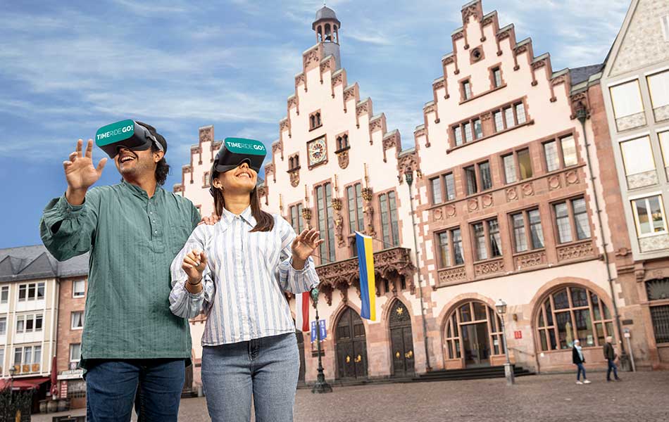 Zwei Personen mit VR-Brillen stehen lächelnd auf dem Römerberg. Im Hintergrund das Rathaus Römer.