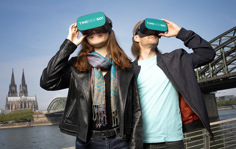 Paar mit VR-Headsets genießt virtuelle Aussicht auf Kölner Dom und Rhein.