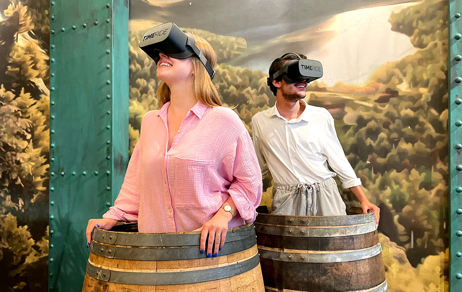 Ein Paar steht mit Virtual Reality Headsets Holzfässern und blicken lachend umher.